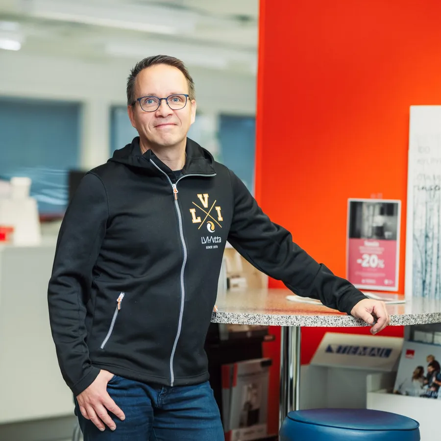 Hymyilevä mies seisoo toimistoympäristössä punaisen seinän vieressä, pukeutuneena mustaan LVI-logolla varustettuun huppariin.