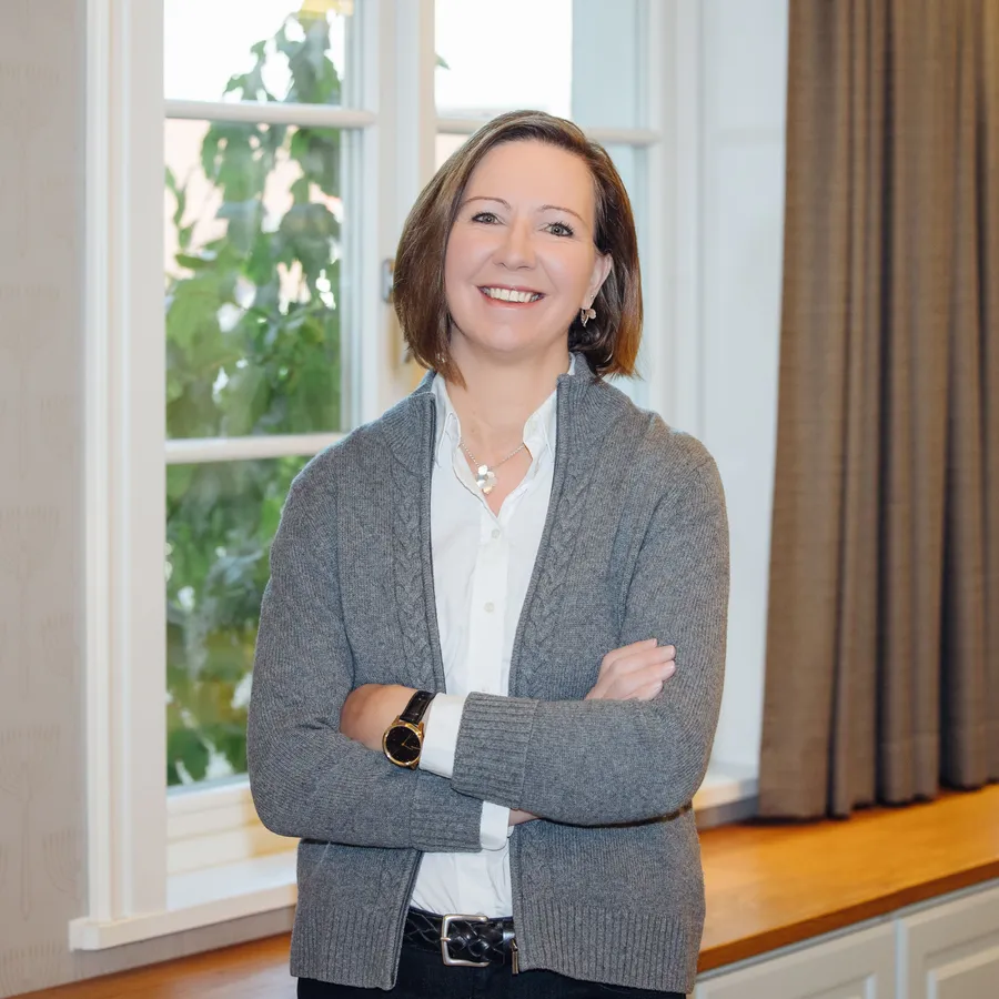 Hymyilevä toimitusjohtaja seisoo valoisassa huoneessa ikkunan äärellä kädet ristissä.