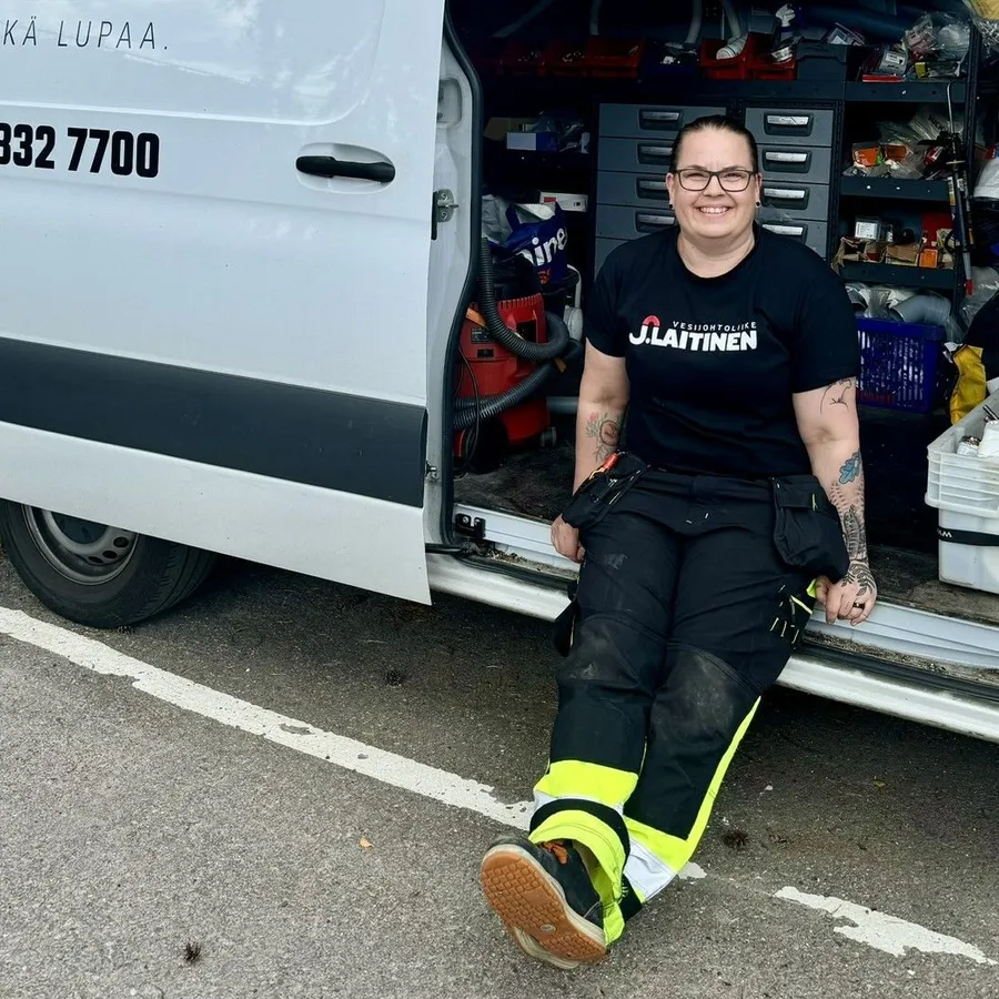 Hymyilevä asentaja istuu työauton avoimella sivuovella työkalujen ja tarvikkeiden keskellä ulkona parkkipaikalla.