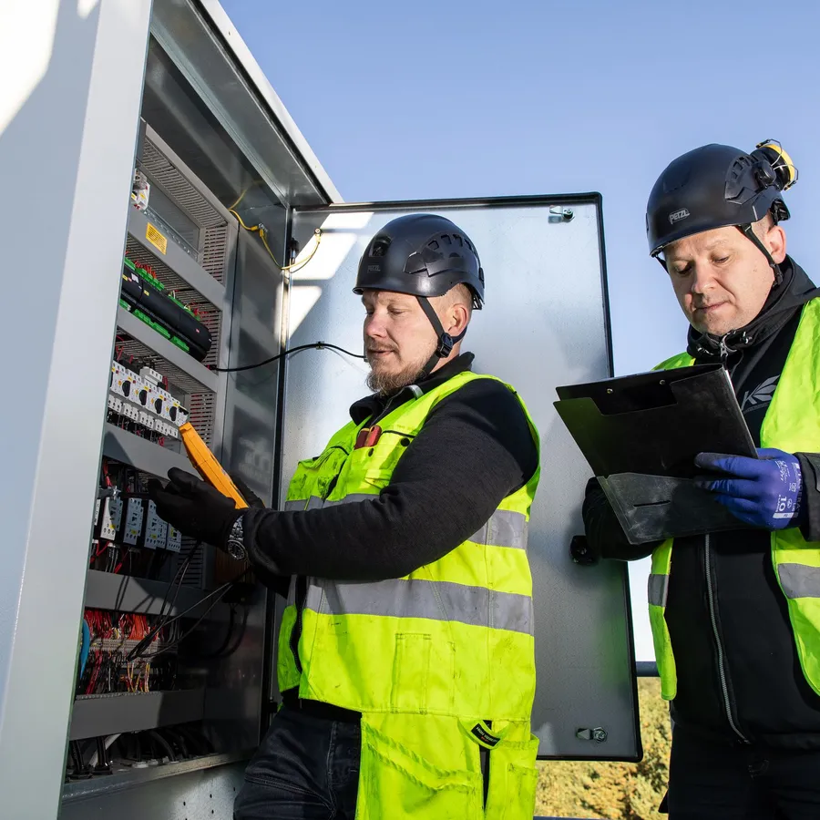 Kaksi asentajaa tarkistaa sähkökaapin mittauksia ja dokumentoi tuloksia ulkona, päällään kypärät ja huomioliivit.