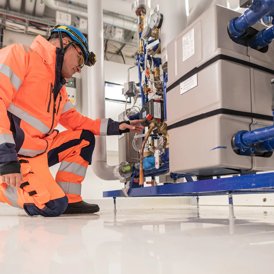 Asentaja tarkastaa teknisen laitteen asetuksia talotekniikkatilan sisällä. Hänellä on päällään kirkas oranssi suojavaatetus ja kypärä.
