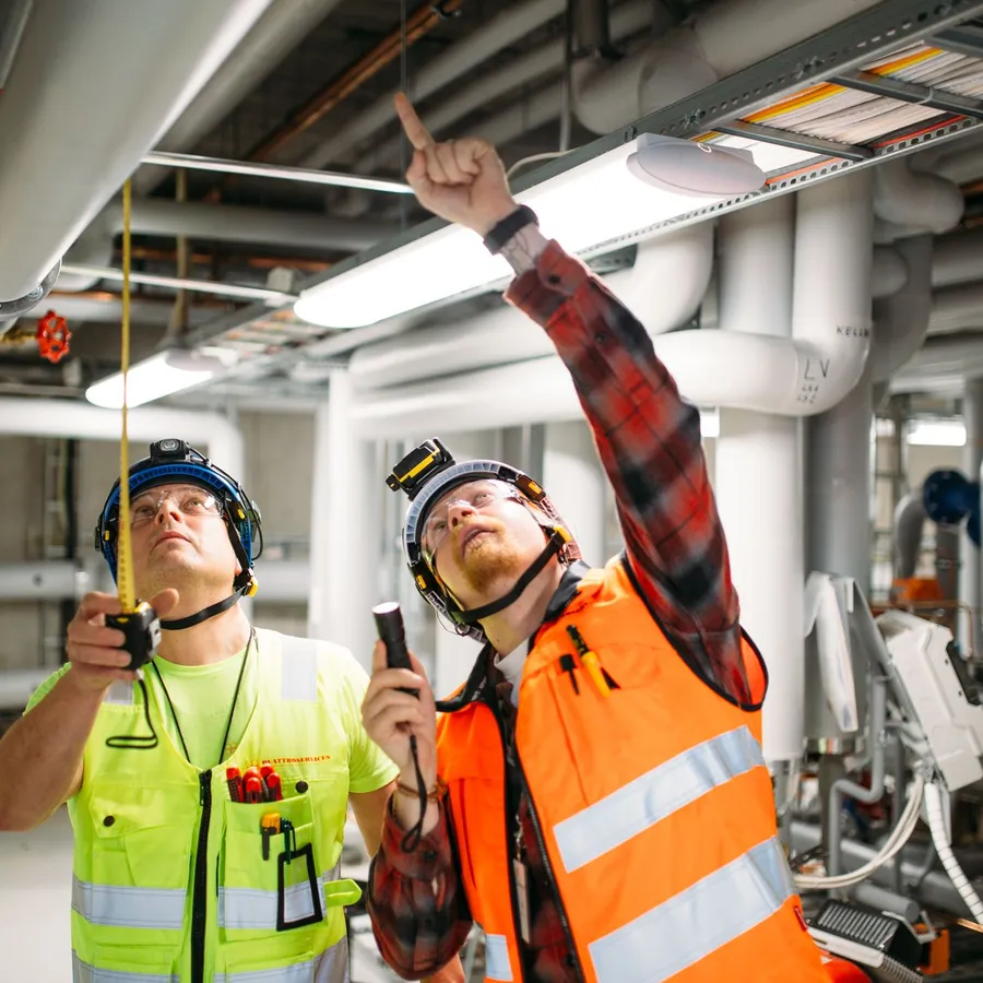 Kaksi talotekniikka-asentajaa tarkastaa ilmanvaihtokanavia ja putkistoja teollisuusrakennuksen konehuoneessa.